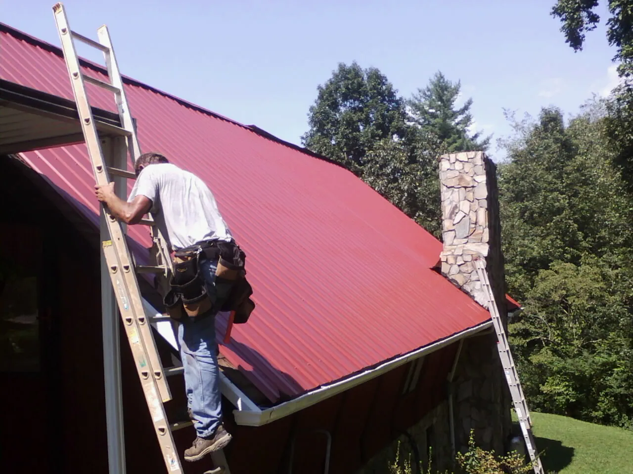 Roofer on a ladder beside a red metal roof and stone chimney — Soffit & Fascia Repair by Roofing Brothers in Becker